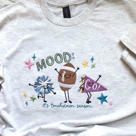 Close-up of colorful embroidered football design featuring pom pom, football and GO banner with stars with the words:  Mood: its football season.  Design is stitched on a Gildan Ash color tee.