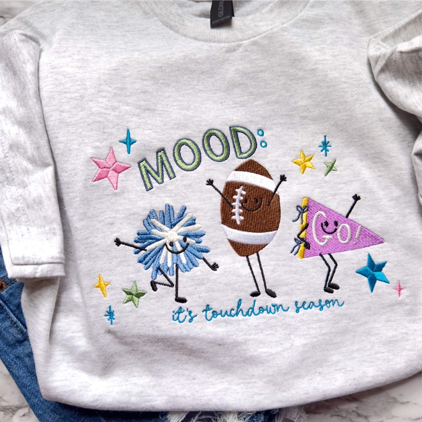 Cute embroidered Gildan football shirt in the color Ash with Mood: It’s Touchdown Season design styled with denim shorts.