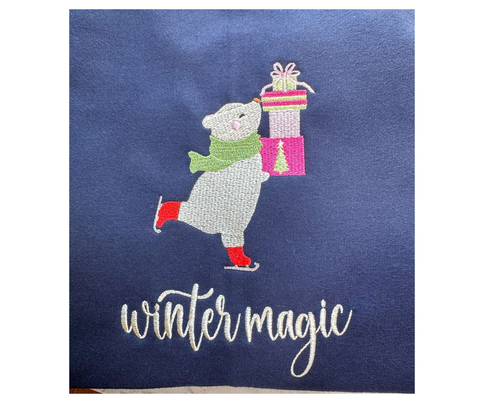 Very cute gildan softstyle Navy sweatshirt with an embroidered design of a polar bear skating while holding Christmas gifts with the words "winter magic".