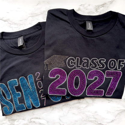 Image of two Senior rhinestone shirts.  One says Class of 2027 with Electric Purple and Crystal lettering and graduation cap in black diamond rhinestones.  The other says Senior 2027 in Capri, chrome and crystal AB rhinestones.  Designs are offered on either a Bella Canvas or Gildan Softstyle black T-shirt.