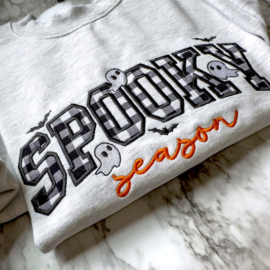 Close up view of a Spooky Season Halloween embroidered design on a Gildan softstyle Ash Gray sweatshirt with checkerboard appliqué, orange script lettering, and cute ghosts and bats.