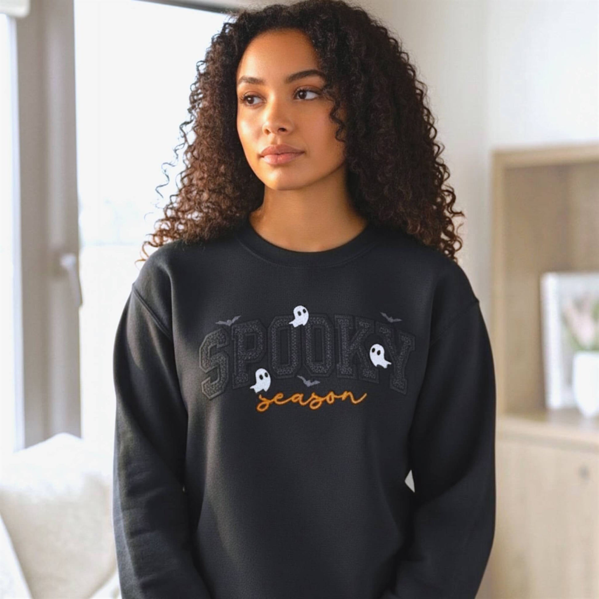 Woman wearing a Gildan Softstyle black Spooky Season Halloween embroidered sweatshirt with black glitter HTV applique, orange script lettering, ghosts and bats.