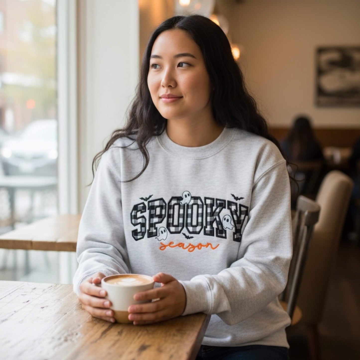 Woman wearing a Gildan softstyle Ash gray Spooky Season Halloween embroidered sweatshirt with checkerboard appliqué, orange script lettering, ghosts and bats.
