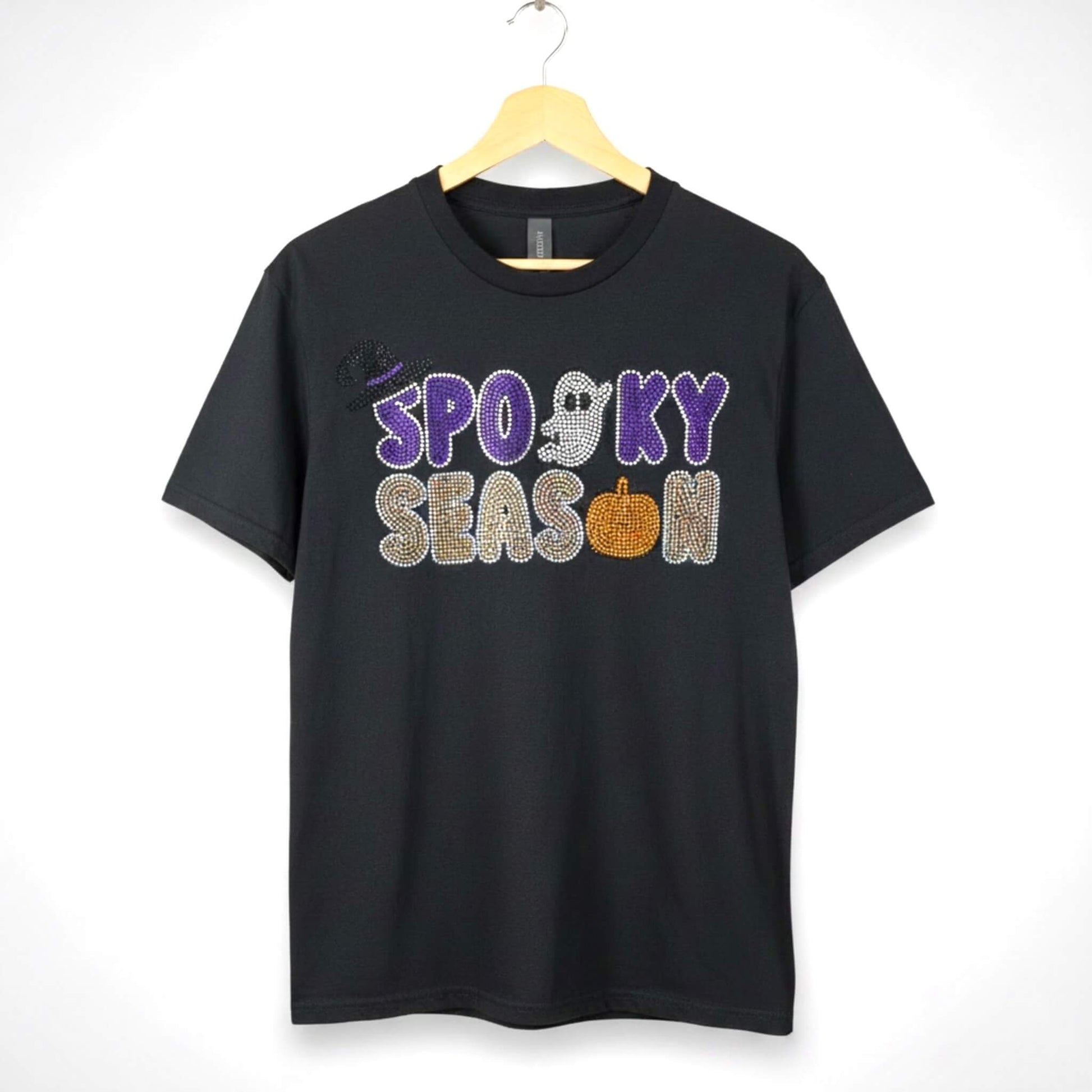 A black short-sleeve t-shirt hanging on a wooden hanger against a white background, featuring a 'Spooky Season' rhinestone design with Halloween themed accents such as a ghost, pumpkin and witch hat.