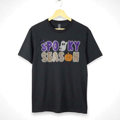 A black short-sleeve t-shirt hanging on a wooden hanger against a white background, featuring a 'Spooky Season' rhinestone design with Halloween themed accents such as a ghost, pumpkin and witch hat.