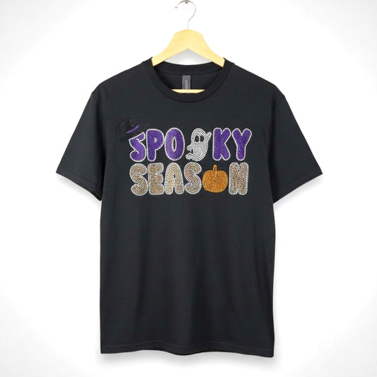 A black short-sleeve t-shirt hanging on a wooden hanger against a white background, featuring a 'Spooky Season' rhinestone design with Halloween themed accents such as a ghost, pumpkin and witch hat.