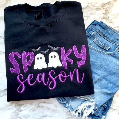 Black Spooky Season Halloween Gildan Softstyle sweatshirt with purple glitter HTV in the word "Spooky" and white glitter inside of the ghosts which replaces the Os in the word Spooky, puff 3D embroidery for the word "Season" and gray bats.  Styled with a pair of jeans.
