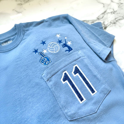 Close-up of embroidered volleyball design and personalized jersey number in blue glitter HTV on Comfort Colors Blue Jean tee with permanently closed pocket.