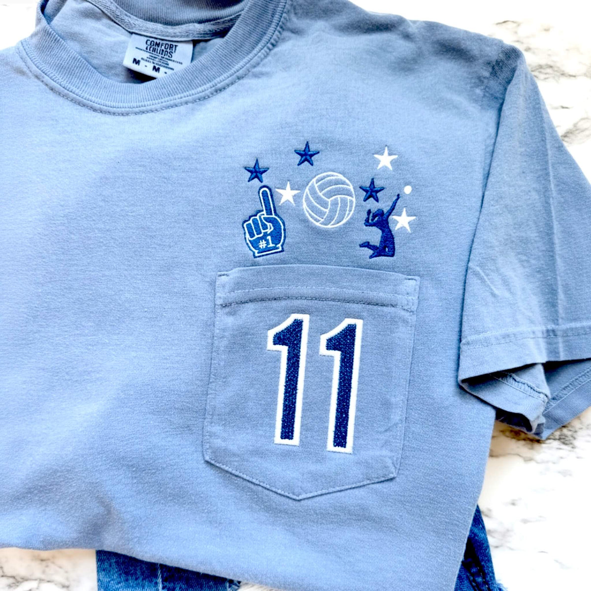 Personalized Comfort Colors Blue Jean volleyball pocket tee with embroidered volleyball design, custom jersey number in blue glitter HTV, and permanently closed pocket.