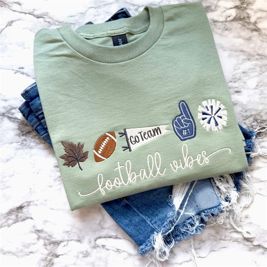 Sage green Gildan Softstyle T-shirt folded to show an embroidered football themed design with fall and game day icons and the word "football vibes" in a script font.  Shirt has been paired with jean shorts for display purposes.