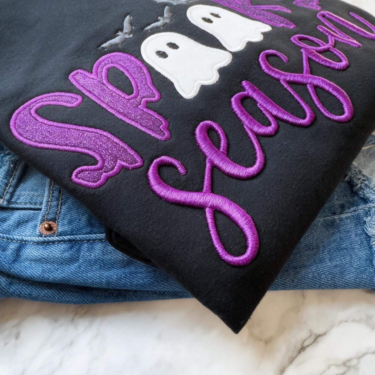 Close up view of a black Spooky Season Halloween Gildan Softstyle sweatshirt with purple glitter HTV in the word "Spooky" and white glitter inside of the ghosts which replaces the Os in the word Spooky, puff 3D embroidery for the word "Season" and gray bats.