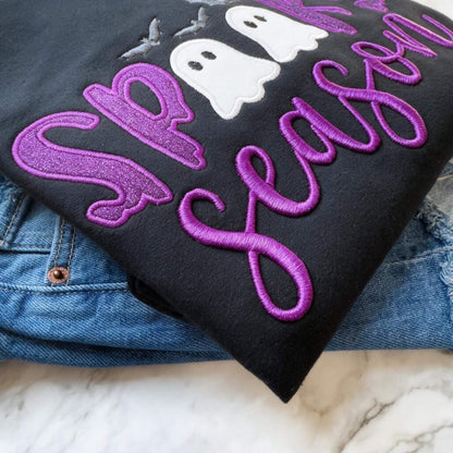 Close up view of a black Spooky Season Halloween Gildan Softstyle sweatshirt with purple glitter HTV in the word "Spooky" and white glitter inside of the ghosts which replaces the Os in the word Spooky, puff 3D embroidery for the word "Season" and gray bats.
