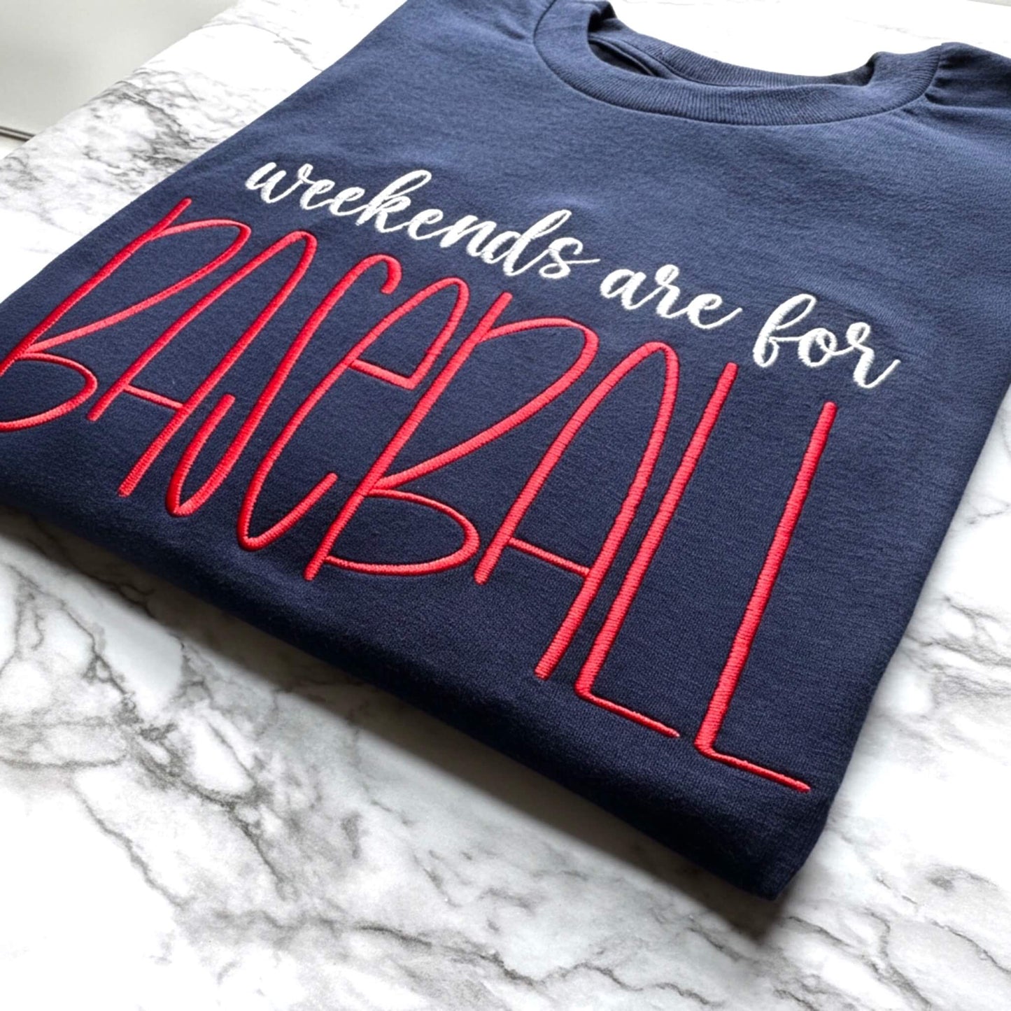 Close up look at a Gildan Navy Embroidered T-Shirt with the words "weekends are for BASEBALL" in white and bright red thread.